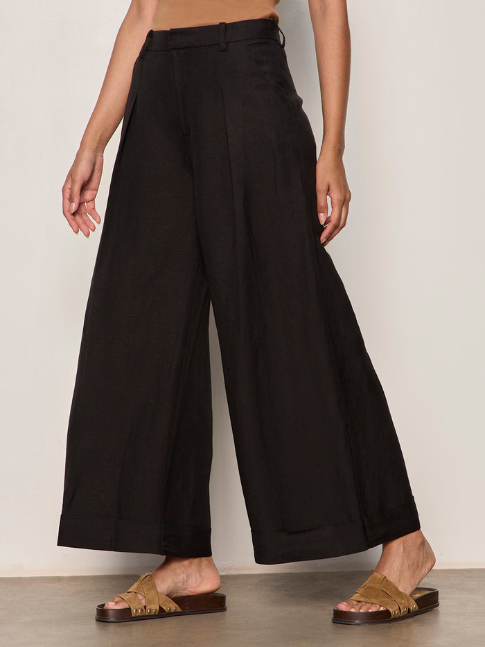 A person stands against a neutral wall wearing Sanctuary Clothing's terrace linen pant in black, paired with a tan top and tan slide sandals. Their arms are relaxed at their sides and only their body from the shoulders down is visible.