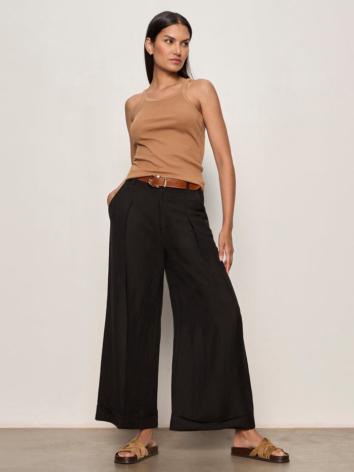 A woman stands against a plain light wall wearing the Sanctuary Clothing terrace linen pant in black, paired with a tan sleeveless top, brown belt, and tan slide sandals. She has straight dark hair and poses with one hand in her pocket.