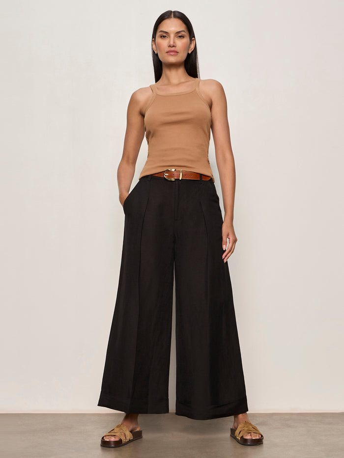 A woman with straight, dark hair stands against a plain light wall wearing Sanctuary Clothing's terrace linen pant in black, styled with a tan sleeveless top, brown belt, and gold sandals. Her hands rest in her pockets.