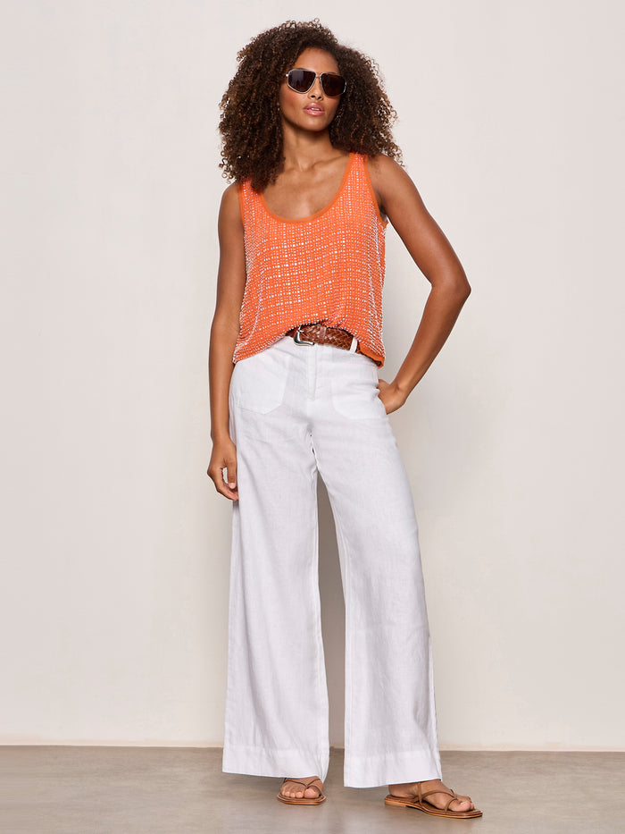 A woman models the becoming bead shell hibiscus orange sleeveless top by Sanctuary Clothing, paired with white wide-leg pants, a brown belt, brown sandals, and sunglasses against a plain light background.