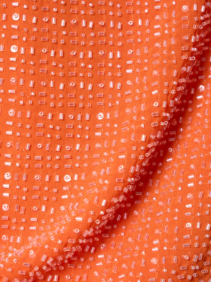 Close-up of the becoming bead shell hibiscus orange by Sanctuary Clothing, featuring orange fabric adorned with clear sequins and beads in a scattered grid pattern for a sparkling, textured look. Slight folds add depth and shadow.