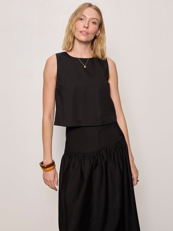 A woman with straight blonde hair wears the desired summer crop shell in black by Sanctuary Clothing, paired with a matching black skirt and accessorized with a gold necklace, hoop earrings, and a brown bangle against a plain light backdrop.