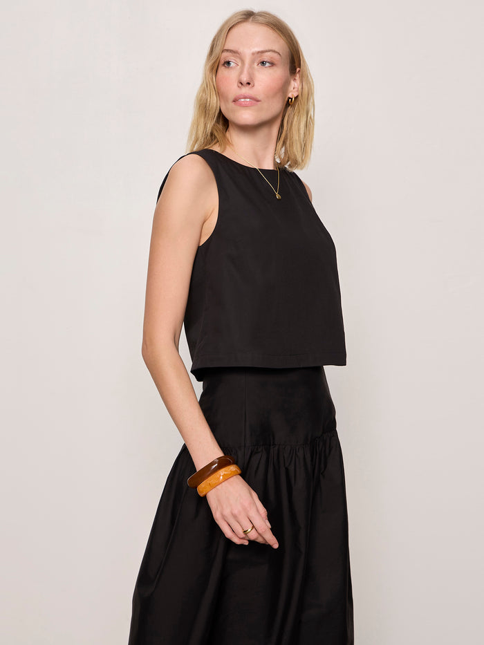 A woman with blonde hair wears Sanctuary Clothing’s “the desired summer crop shell” in black, paired with a matching high-waisted skirt. She accessorizes with gold earrings, a simple necklace, and amber bangles, standing against a plain light background.