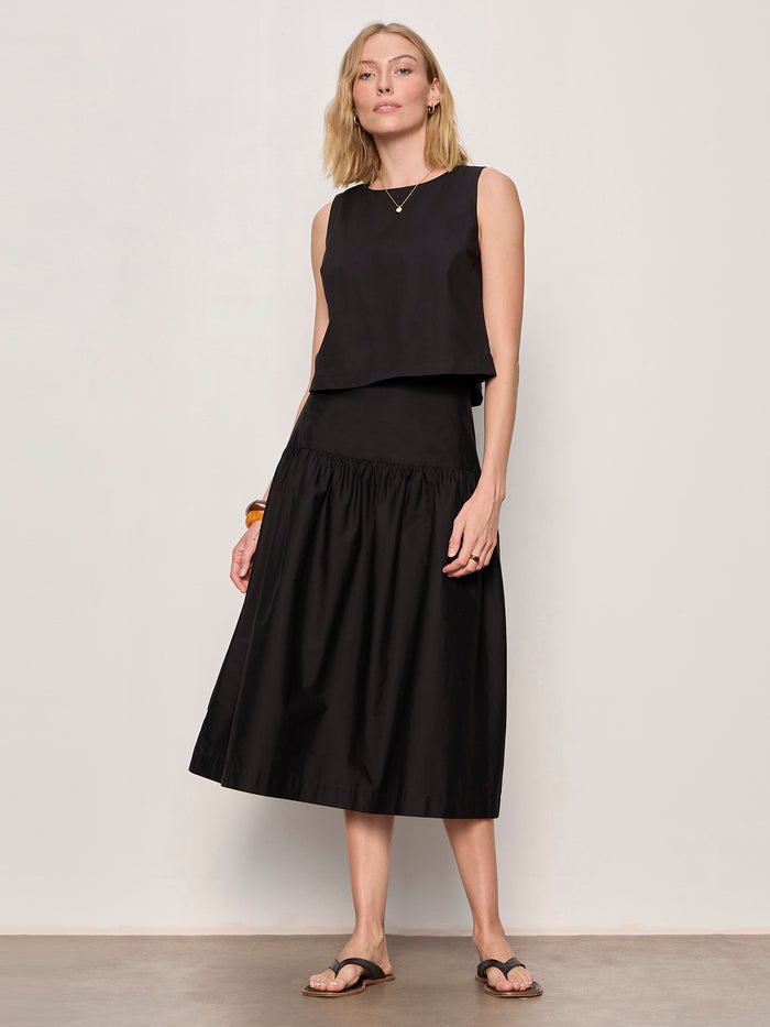 A woman poses confidently against a light wall, wearing Sanctuary Clothing's the desired summer crop shell in black with a black midi skirt, sandals, gold necklace, and chunky bracelet.