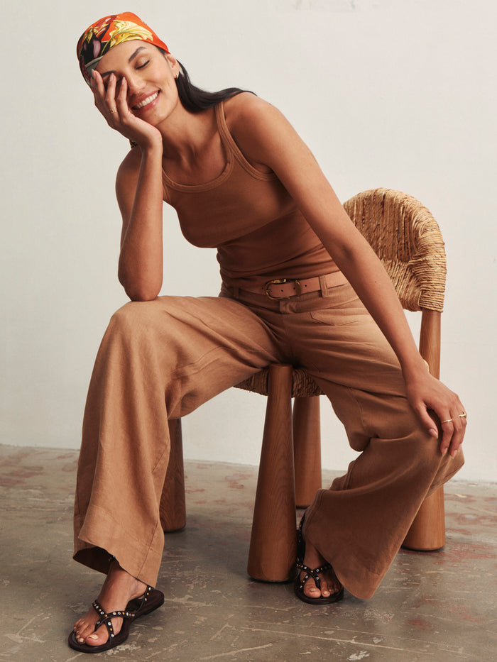 A smiling woman wears the linen marine wide leg toasted coconut pants by Sanctuary Clothing, paired with a brown tank top and black sandals. She sits relaxed on a wooden chair, her colorful headscarf adding a cheerful touch.