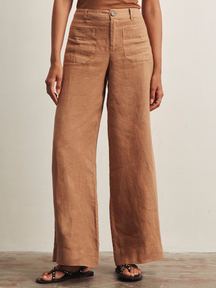 A person wearing Sanctuary Clothing’s linen marine wide leg toasted coconut pants, high-waisted with large front pockets and a button closure, paired with black studded sandals, stands against a light-colored wall.