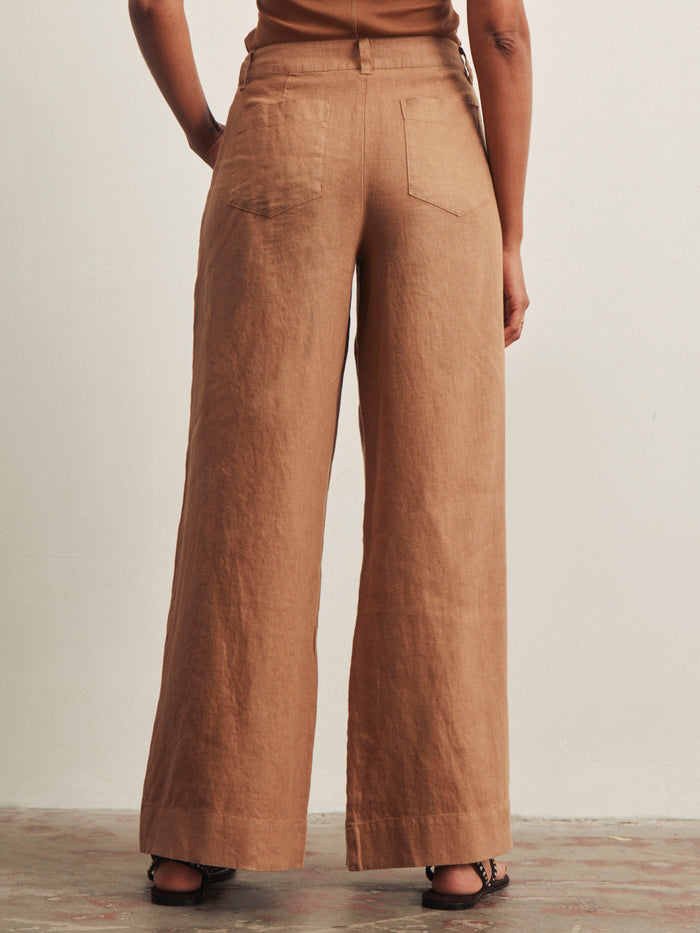 A person stands facing away, wearing Sanctuary Clothing's the linen marine wide leg toasted coconut pants with two back pockets, paired with sandals. The background is a plain light-colored wall.