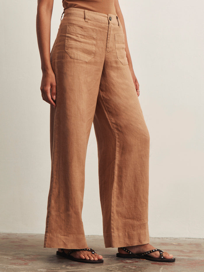 A person wears Sanctuary Clothing's the linen marine wide leg toasted coconut pants with large front pockets, paired with a beige top and black studded sandals, standing against a neutral background.