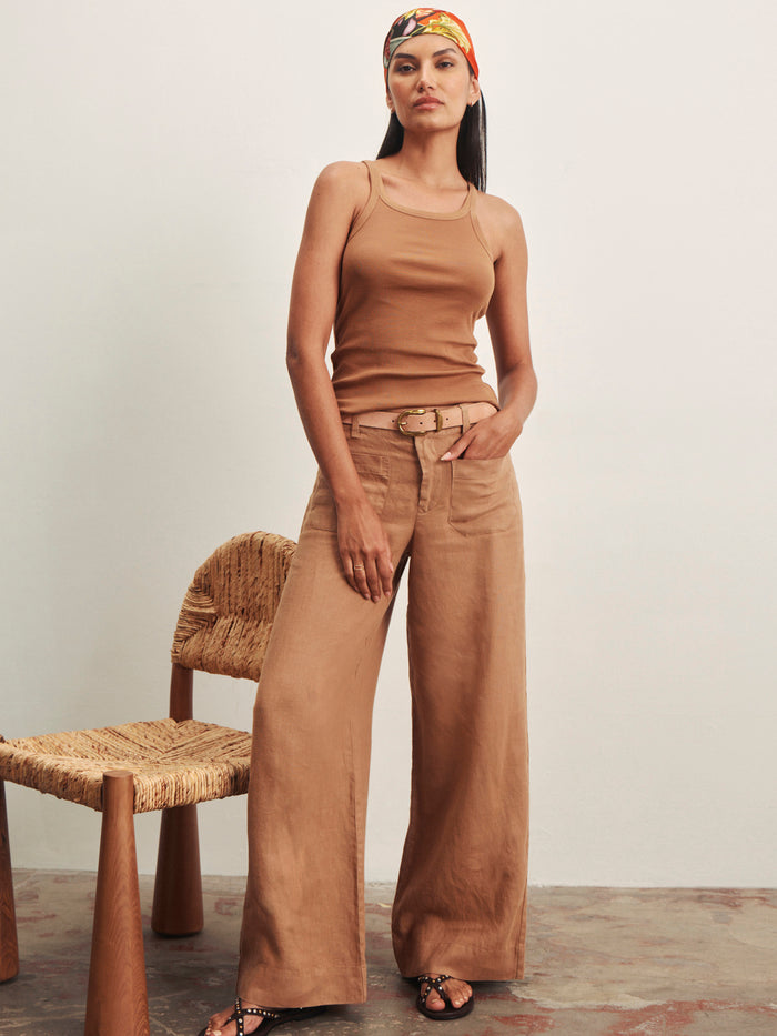 A woman stands confidently indoors with one hand in her pocket, wearing Sanctuary Clothing's the linen marine wide leg toasted coconut pants, a tan tank top, sandals, and a colorful headscarf beside a woven chair.