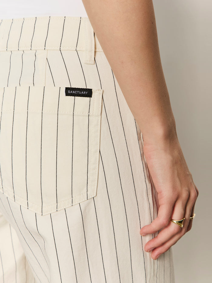 A close-up of someone wearing Sanctuary Clothing's the marine jean crop bleached white pinstripe petite jeans, featuring thin black stripes and a branded back pocket label, with a gold ring on their finger.