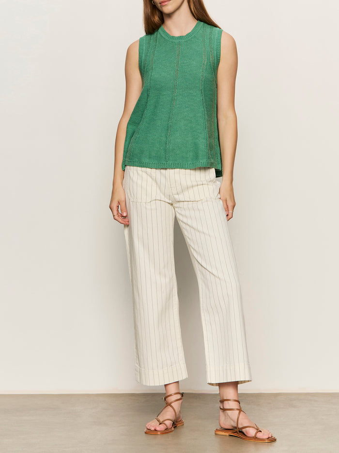 A person stands against a plain background wearing the Sanctuary Clothing marine jean crop in bleached white pinstripe (petite sizing), a sleeveless green top, and brown strappy sandals. Their hair is down, and their arms are relaxed at their sides.