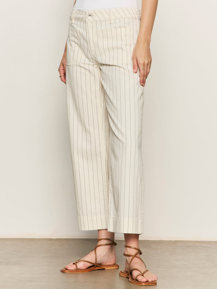 A person stands indoors wearing Sanctuary Clothing's The Marine Jean in Bleached White Pinstripe. Only the lower half of the body is visible, showing ankle-length pants with thin vertical stripes, a white top, and brown flat sandals.