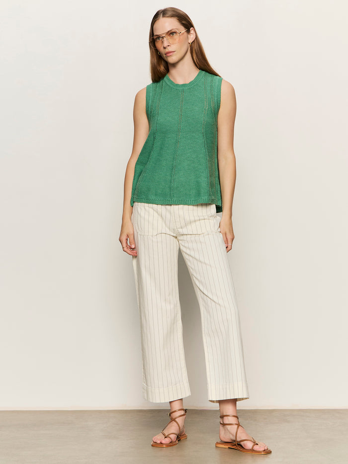 A woman with long straight hair and clear eyeglasses wears a sleeveless green top, brown strappy sandals, and Sanctuary Clothing's the marine jean bleached white pinstripe against a light-colored wall. She has a neutral expression.