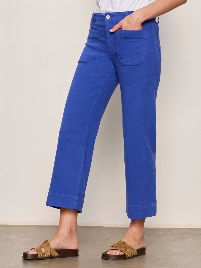 A person models Sanctuary Clothing’s “the marine luminous blue” cropped jeans with large front pockets, paired with a white tucked-in shirt and tan slide sandals against a light background, one hand casually in their pocket.