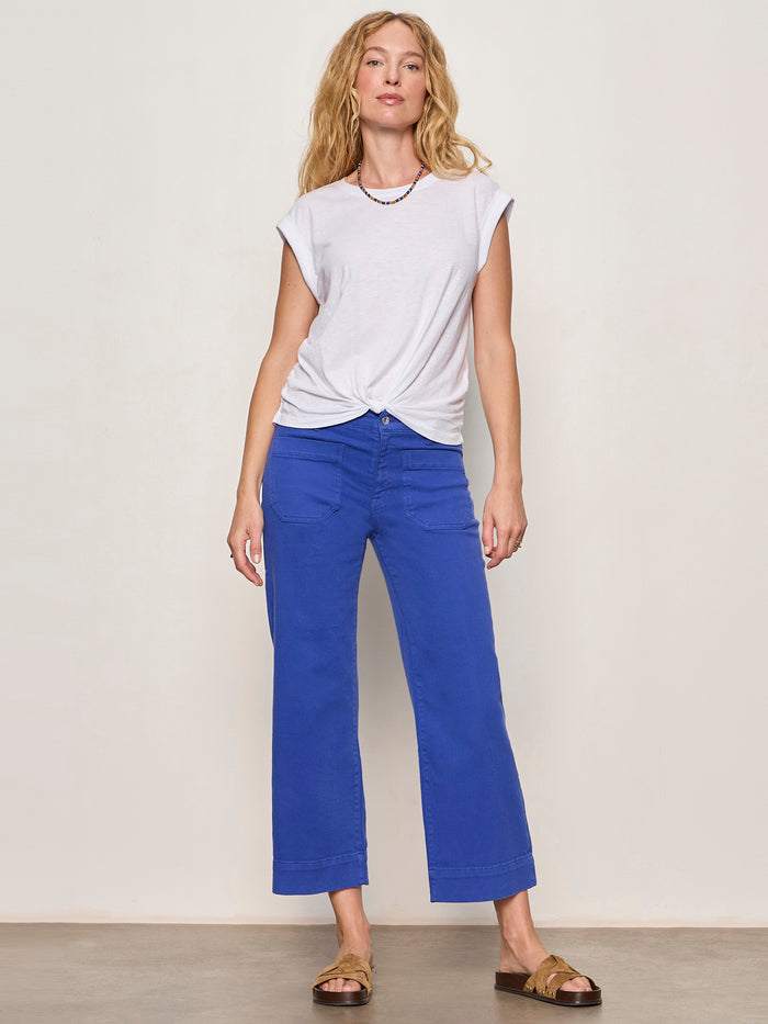 A woman with long blonde hair poses relaxed against a plain background, wearing Sanctuary Clothing’s the marine luminous blue wide-leg pants, a white knotted t-shirt, a colorful beaded necklace, and brown sandals.