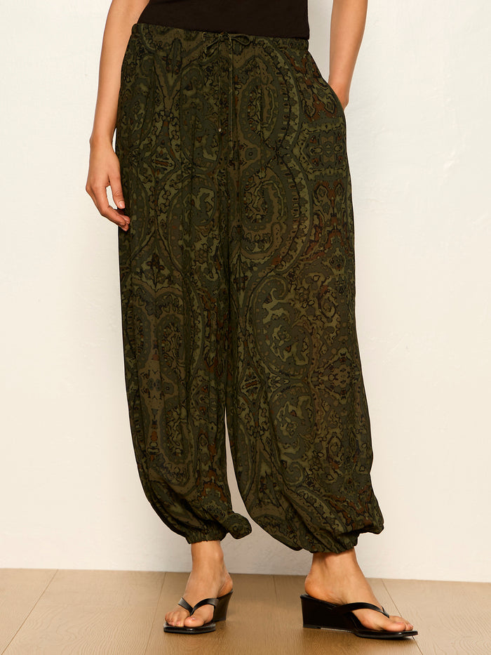 A person wears Sanctuary Clothing's the moment volume pant in midnight moss, featuring a loose fit and elastic cuffs, paired with a black top and black sandals, standing against a light wall on a wooden floor.