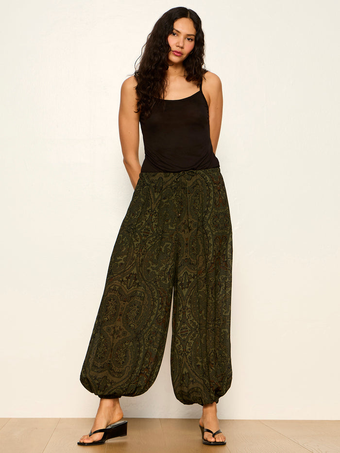A woman with long, wavy hair wears the Sanctuary Clothing "the moment volume pant" in midnight moss, a black sleeveless top, and black sandals. She stands against a plain light-colored wall with a relaxed expression.