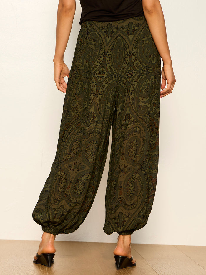 A person wearing Sanctuary Clothing’s the moment volume pant in midnight moss, featuring a loose fit and elastic cuffs, paired with black heeled sandals, stands against a light background, photographed from the back.