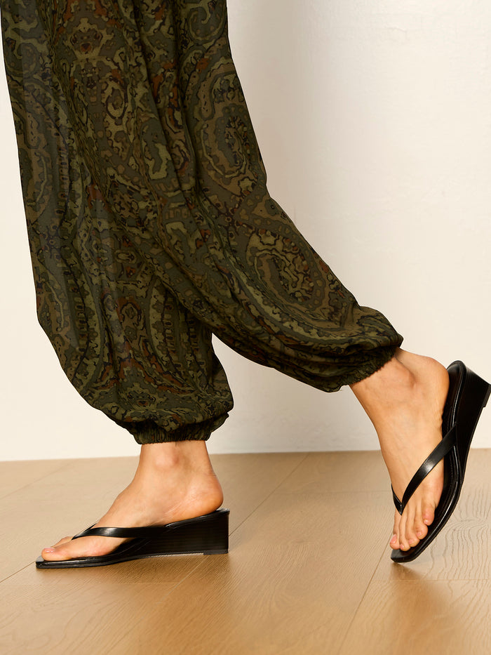 Someone wears the Sanctuary Clothing "the moment volume pant" in midnight moss, styled with black wedge sandals, walking on a light wooden floor against a plain off-white wall.