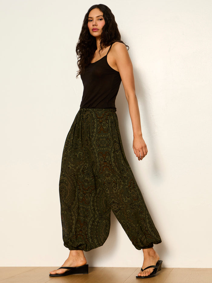 A woman with long dark hair wears the moment volume pant in midnight moss by Sanctuary Clothing, paired with a black tank top and sandals, standing against a plain light wall and wooden floor.