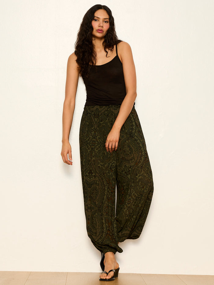 A woman with long wavy hair wears a black tank top, black sandals, and Sanctuary Clothing's the moment volume pant in midnight moss. She stands against a plain light wall, one arm relaxed and the other slightly bent.