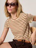 A woman with blonde hair and dark sunglasses sits on a chair wearing Sanctuary Clothing's the perfect tee choco banana stripe and brown shorts, with a neutral expression and one hand resting on her hip.