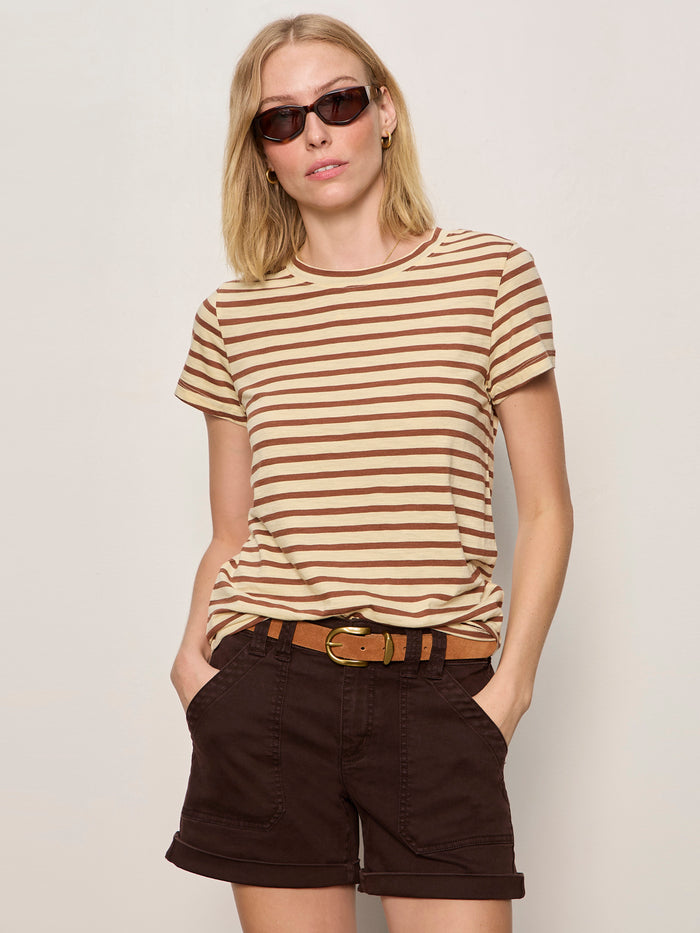 A woman wearing the Sanctuary Clothing "the perfect tee choco banana stripe" stands against a plain light-colored wall, pairing the striped t-shirt with sunglasses and dark brown belted shorts, her hands in her pockets.
