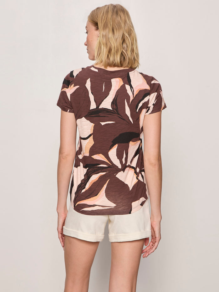 A person with blonde hair, seen from behind, wears the Sanctuary Clothing "the perfect tee desertscape," featuring brown and pink abstract patterns, paired with light shorts against a plain light background.