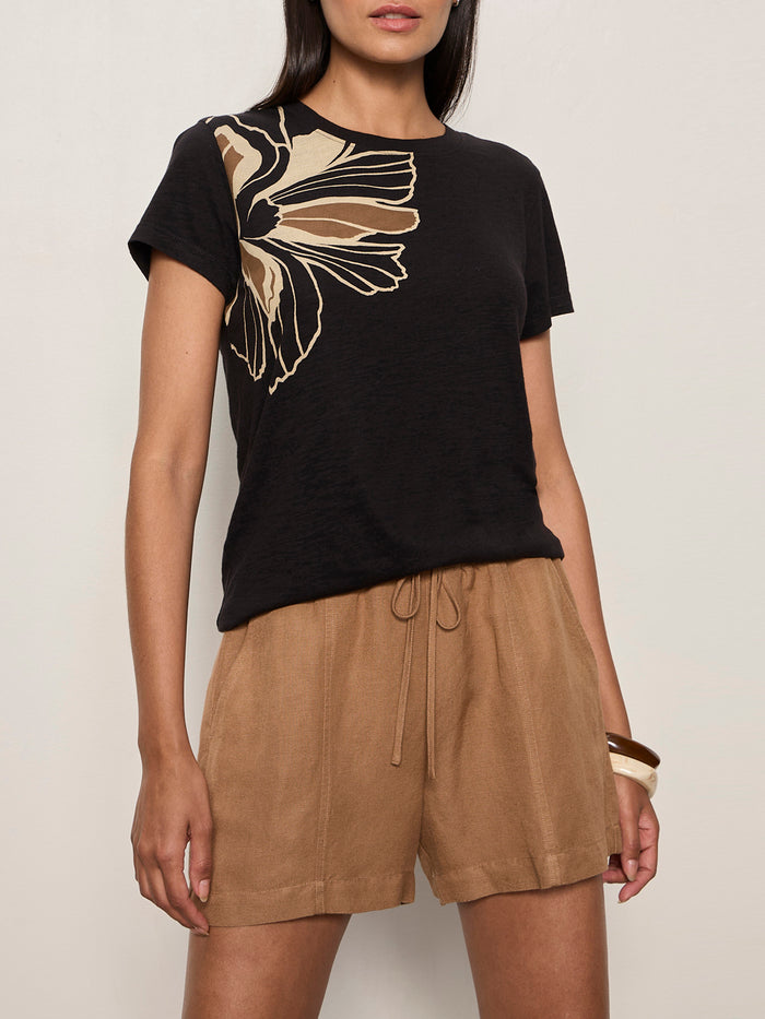 A woman wears the Sanctuary Clothing perfect tee lotus placement, a black top featuring a beige abstract floral design on the shoulder. She pairs it with loose high-waisted tan shorts and a chunky bracelet against a plain light background.
