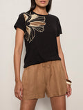 A woman wears the Sanctuary Clothing perfect tee lotus placement, a black top featuring a beige abstract floral design on the shoulder. She pairs it with loose high-waisted tan shorts and a chunky bracelet against a plain light background.