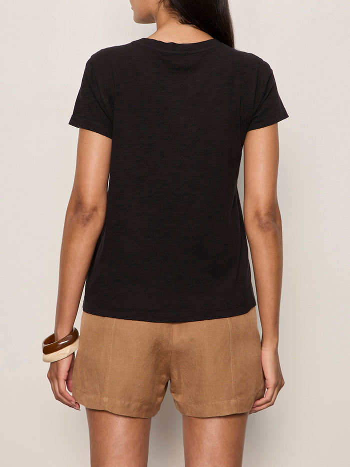 A woman faces away from the camera, wearing Sanctuary Clothing's perfect tee lotus placement in black, paired with tan shorts and two chunky bracelets—one brown, one cream—on her left wrist against a plain off-white wall.