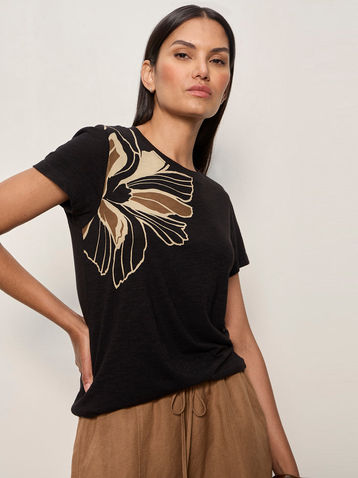 A woman with long, straight dark hair confidently poses in Sanctuary Clothing’s “the perfect tee lotus placement,” a black t-shirt featuring a large beige and tan abstract floral design on one shoulder, paired with light brown pants.