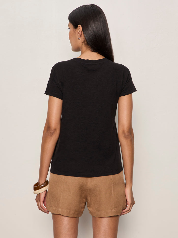 A woman with long dark hair wears the Sanctuary Clothing "the perfect tee lotus placement" black t-shirt, tan shorts, and two brown bangles, standing with her back to the camera against a plain light wall.
