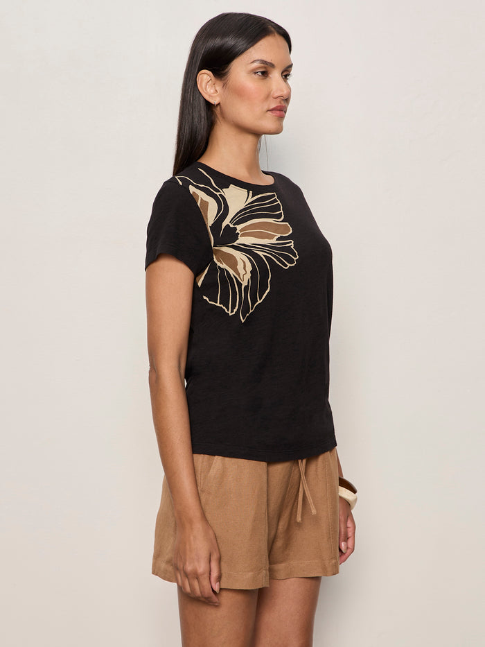 A woman with long dark hair and a neutral expression stands in profile against a plain light wall, wearing Sanctuary Clothing’s “the perfect tee lotus placement” in black with a large tan floral design on the shoulder and tan drawstring shorts.