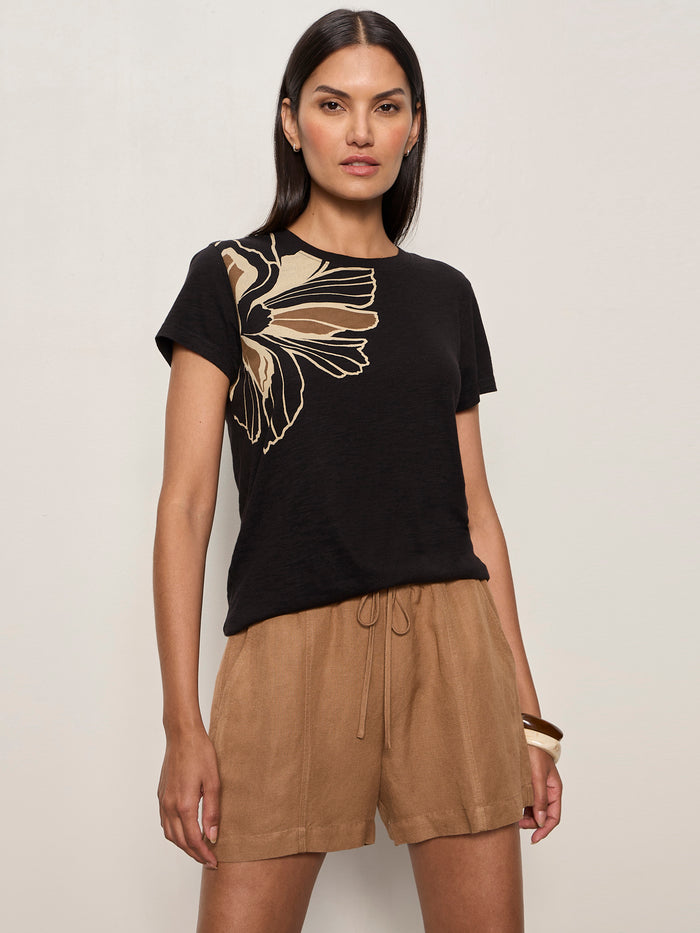 A woman with straight dark hair and a neutral expression models Sanctuary Clothing's "the perfect tee lotus placement," a black tee with a beige floral graphic on the shoulder, paired with loose tan shorts. One hand is in her pocket, set against a light background.