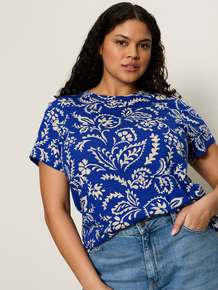 A woman with long curly hair wears the perfect tee portofino extended sizing by Sanctuary Clothing, paired with light blue jeans, standing against a plain light background.