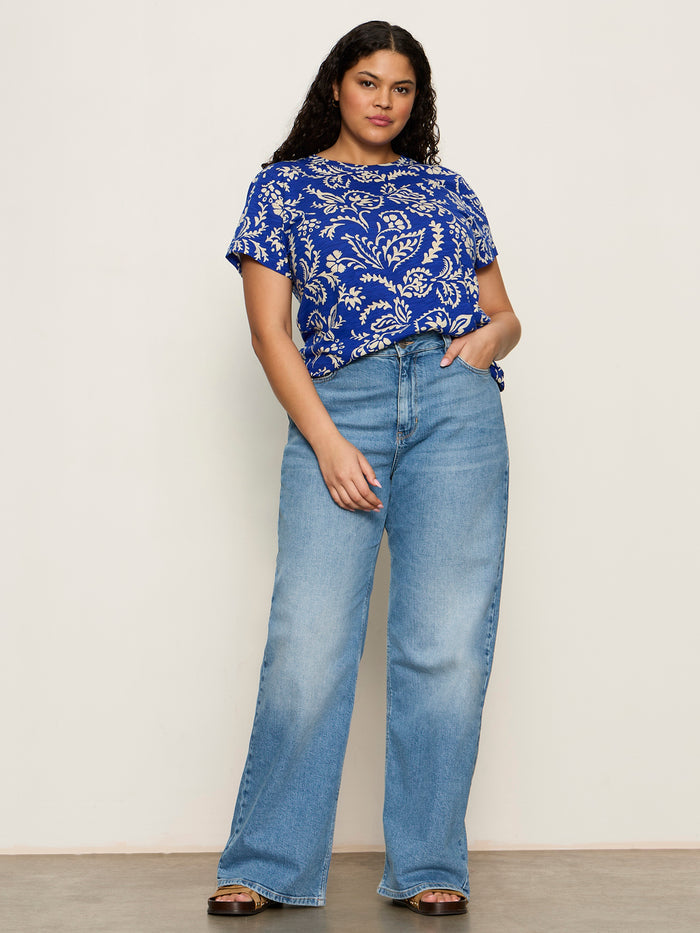 A woman with long, wavy hair and a relaxed expression models Sanctuary Clothing’s "the perfect tee portofino extended sizing" against a plain background, paired with high-waisted wide-leg jeans and brown sandals.