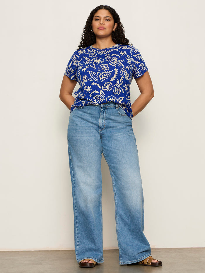 A woman with curly hair and a confident look stands against a plain background, hands in pockets, wearing Sanctuary Clothing's the perfect tee portofino extended sizing—styled with wide-leg light blue jeans and brown sandals.
