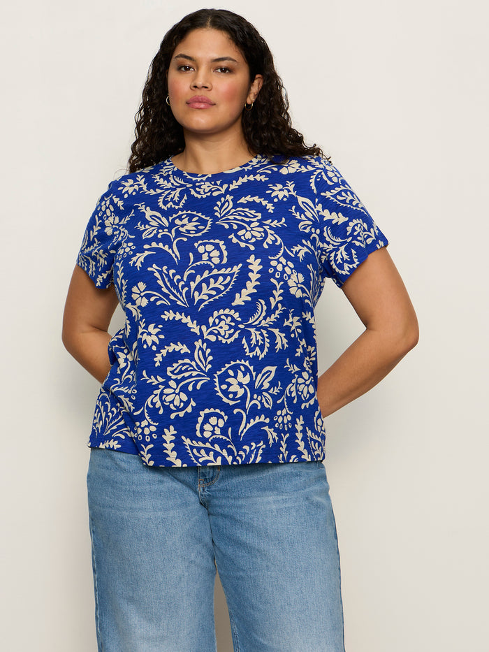A woman with long curly hair wears the Sanctuary Clothing "the perfect tee portofino extended sizing" shirt, featuring blue short sleeves and a white floral pattern, paired with light blue jeans against a plain background.