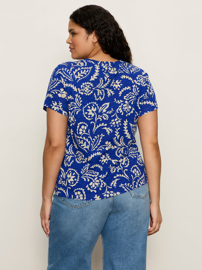 A woman with wavy dark hair, seen from behind against a light background, wears Sanctuary Clothing's the perfect tee portofino extended sizing—a blue short-sleeve shirt with a white floral pattern—and light blue jeans.