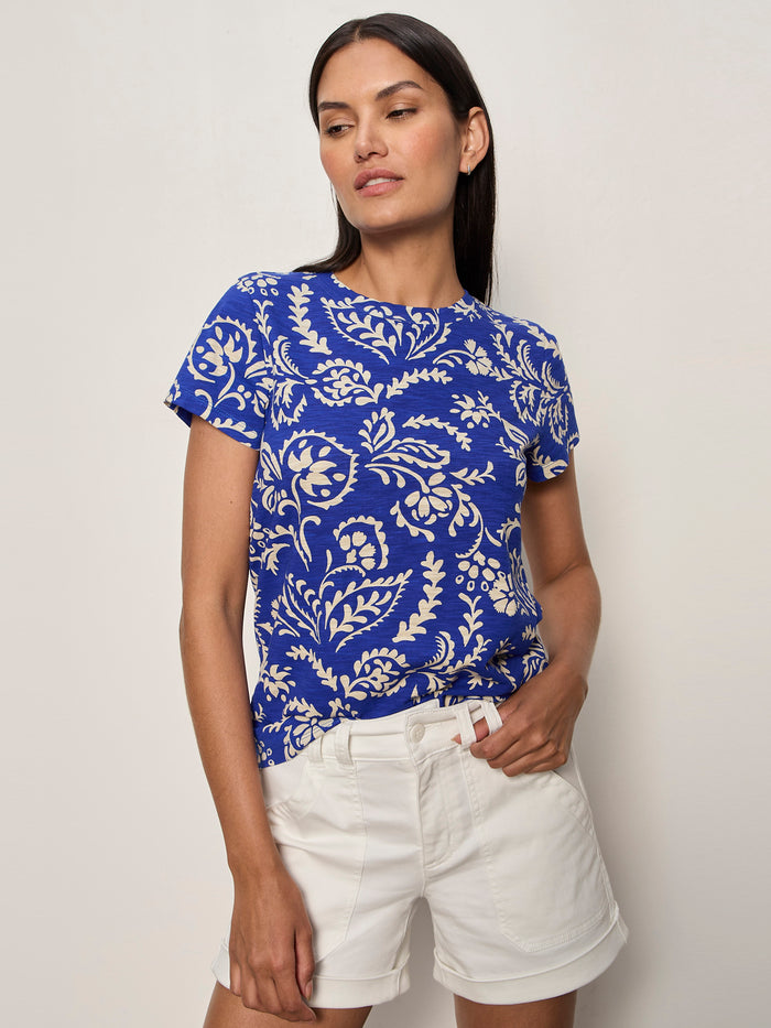 A woman wearing the Sanctuary Clothing “the perfect tee portofino,” a blue short-sleeve shirt with a white floral pattern, and white shorts stands against a light background, looking to the side with one hand in her pocket.