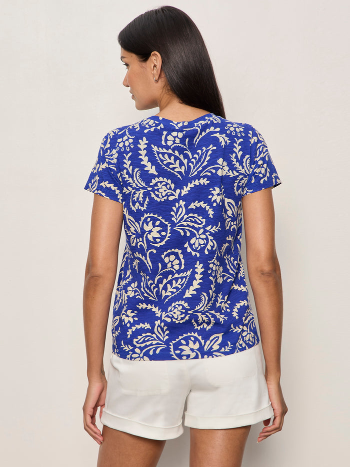 A woman stands facing away against a light background, wearing Sanctuary Clothing's "The Perfect Tee Portofino," a blue short-sleeve shirt with a white leafy pattern, paired with white shorts.
