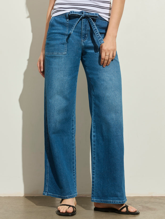 A person wears the Sanctuary Clothing workshop jean utility blue—high-waisted, wide-leg jeans with a matching denim belt and front patch pockets—styled with a striped top and black sandals against a light background.