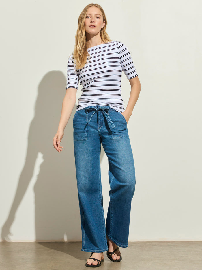 A woman stands against a plain background wearing a black-and-white striped top, Sanctuary Clothing workshop jean utility blue high-waisted wide-leg jeans with a tie belt, and black sandals. One hand is in her pocket.
