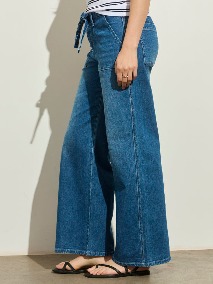 Someone is wearing the Sanctuary Clothing workshop jean utility blue, paired with a striped shirt and black sandals, standing sideways against a light-colored wall.