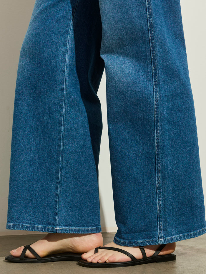 A person wears Sanctuary Clothing's workshop jean utility blue with black strappy flat sandals, standing on a concrete floor near a white wall. Only their lower legs and feet are visible.